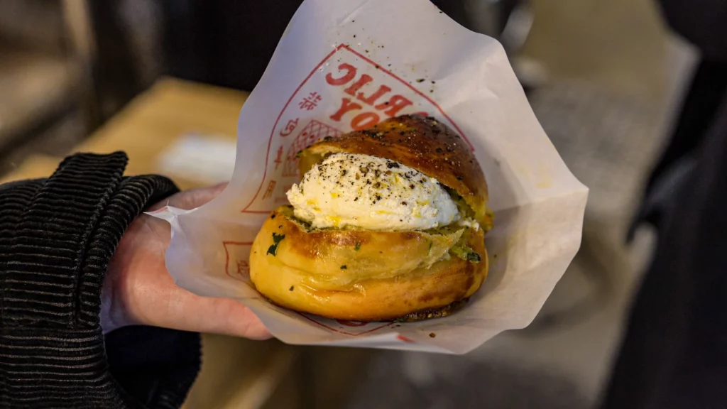 A hand holding a Garlic Boy truffle cream garlic bread in branded paper wrapper, showing the cream cheese filling dusted with black pepper.