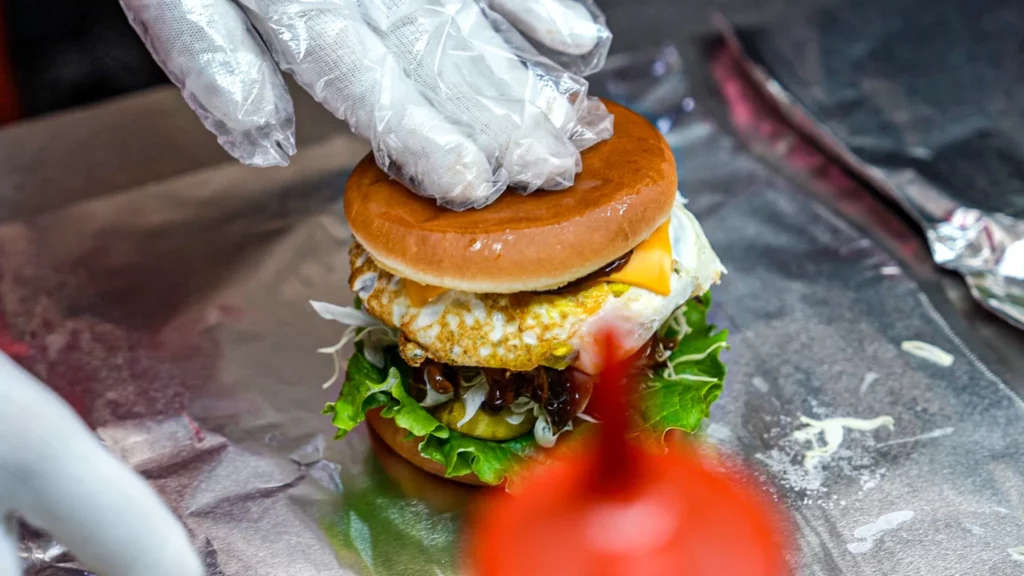 Samdeok Burger vendor pressing the top bun onto a fully stacked bulgogi burger loaded with egg, cheese, cabbage, lettuce, and sauce.
