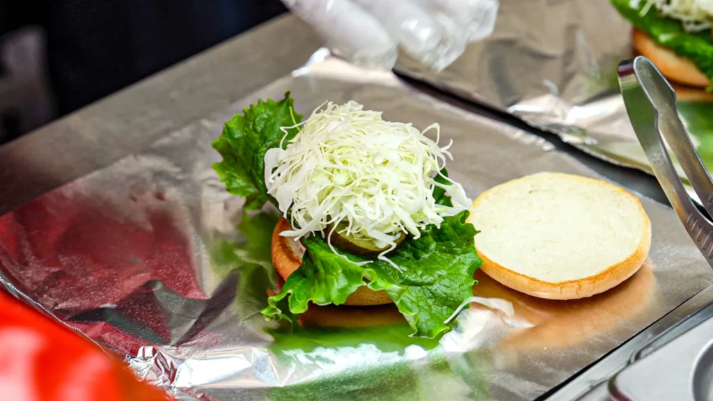 Early assembly stage of a Samdeok Burger showing a generous mound of shredded cabbage over lettuce on the bottom bun, with top bun placed to the side.