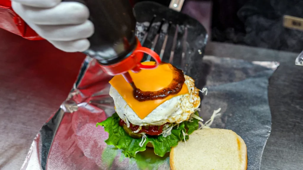 Samdeok Burger vendor squeezing bulgogi sauce from a bottle onto a fried egg and melted cheese slice during burger preparation