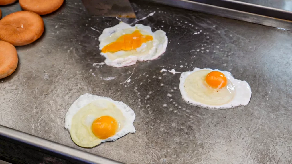 Three sunny-side-up eggs and burger buns cooking simultaneously on a flat iron griddle at Samdeok Burger.