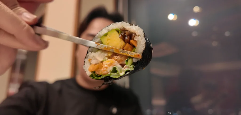 Close-up of Durumi Bunsik kimbap cross-section held by chopsticks showing colorful fillings