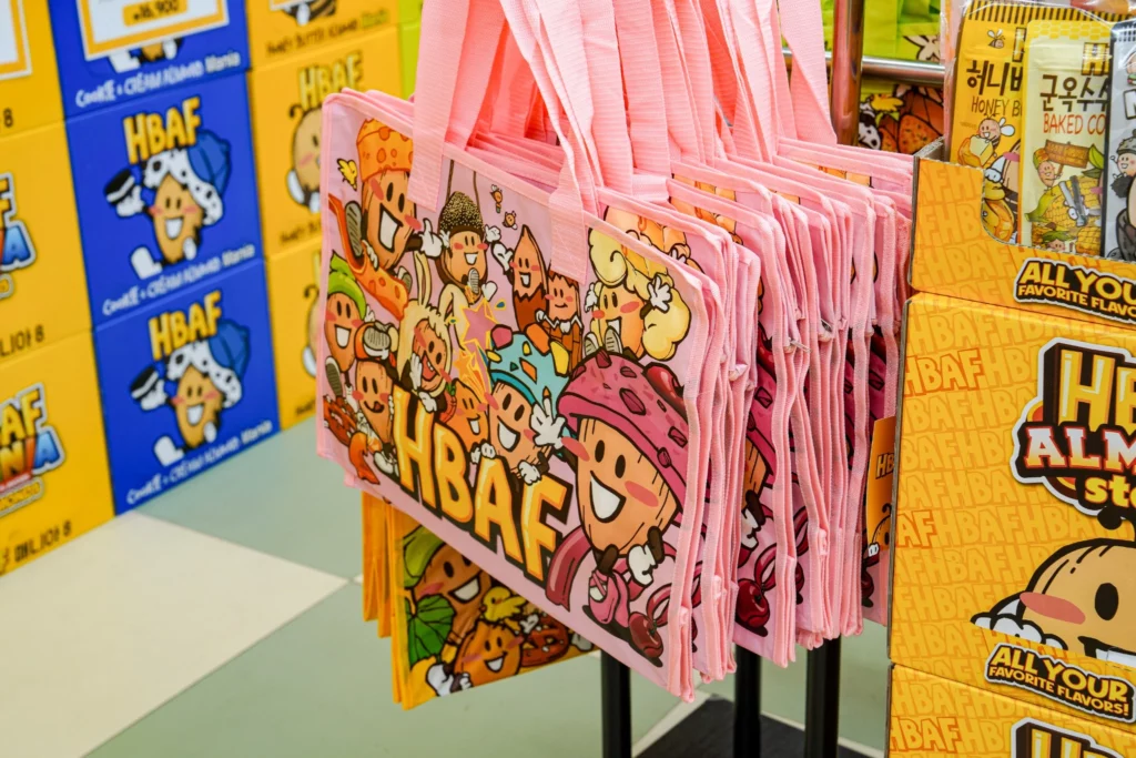 Pink HBAF character-printed reusable tote bags hanging for sale alongside cobalt-blue Cookie & Cream Almond gift boxes.
