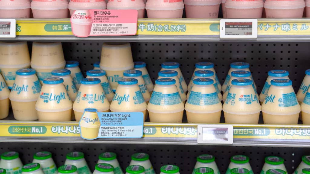 Binggrae Banana Flavored Milk Light bottles displayed with multilingual price tags and strawberry milk on the top shelf.
