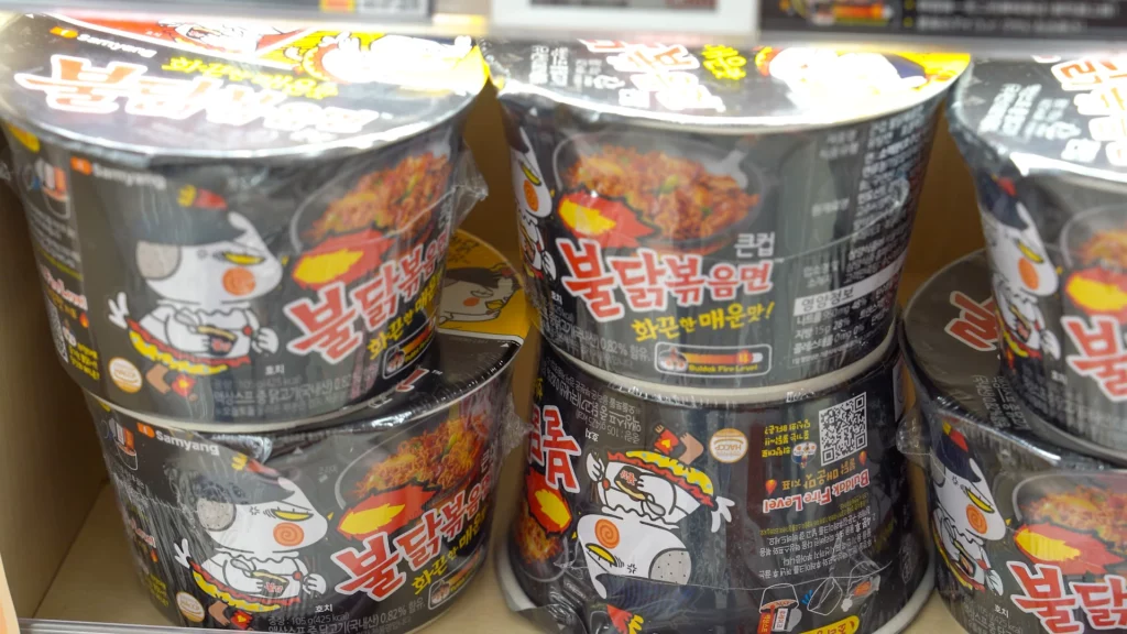 Samyang Buldak Bokkeumyeon spicy chicken cup noodles stacked on a shelf showing the Buldak Fire Level spice indicator.