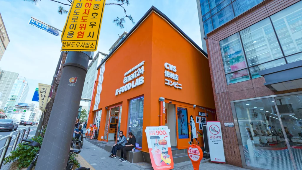 Orange triangular storefront of Emart24 K-Food Lab Myeongdong near Toegye-ro with CVS signage in Korean, Chinese, and Japanese.
