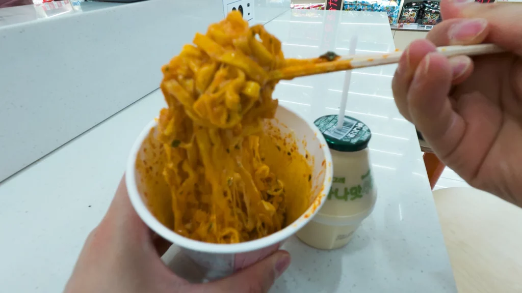 First-person view of freshly cooked Buldak-style ramyeon lifted with chopsticks from a paper bowl beside a Binggrae banana milk.
