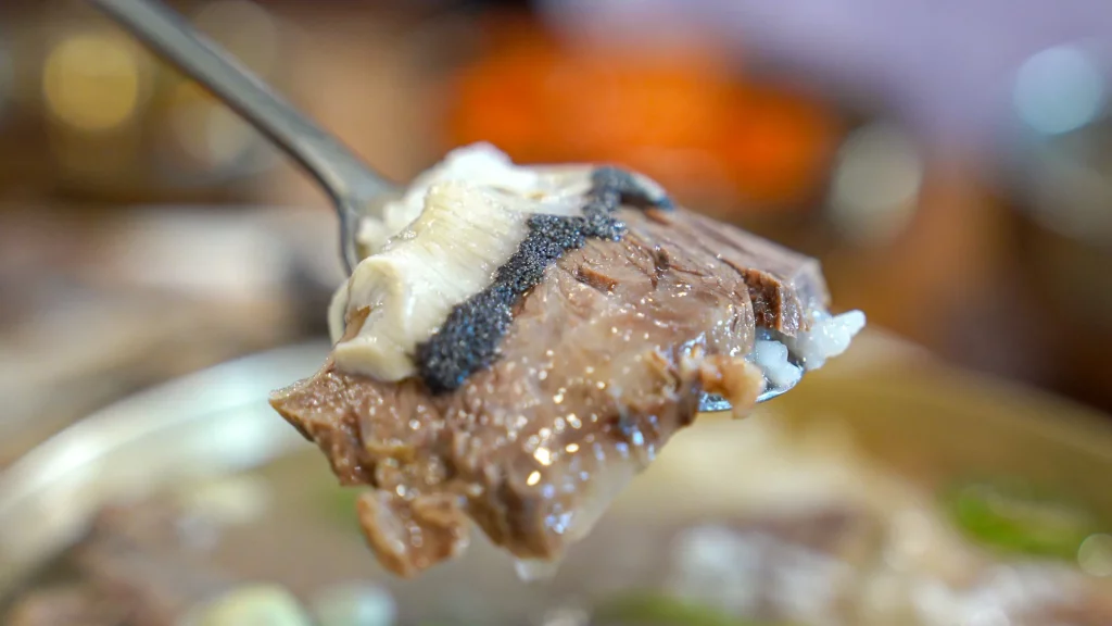 Macro shot of a tender Hanwoo brisket slice and tripe lifted on a spoon over a Hadongkwan gomtang bowl.
