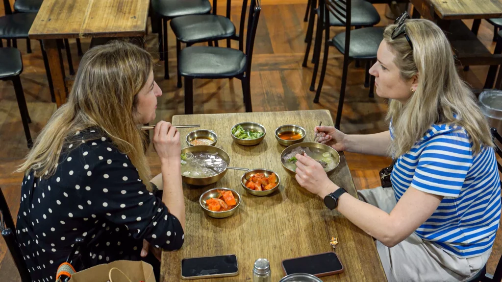 Two foreign tourists sharing bowls of Hadongkwan Myeongdong Hanwoo gomtang at a wooden table with brass side dishes.
