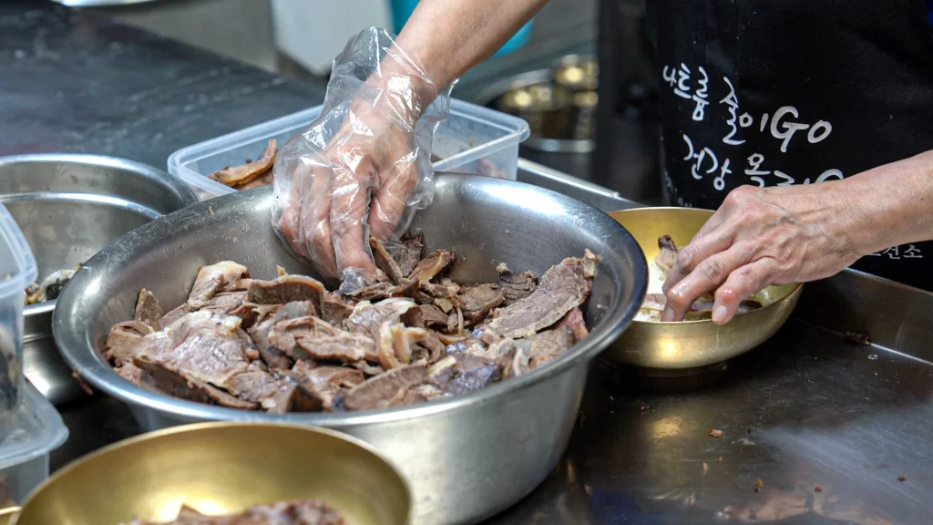 Gloved hands placing sliced Hanwoo brisket onto rice in a brass bowl, the signature plating step at Hadongkwan Myeongdong.