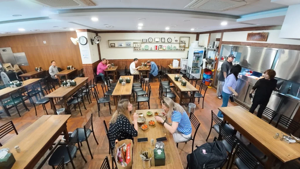 Wide view of Hadongkwan Myeongdong's wooden-floor dining hall with foreign tourists eating gomtang at center tables.