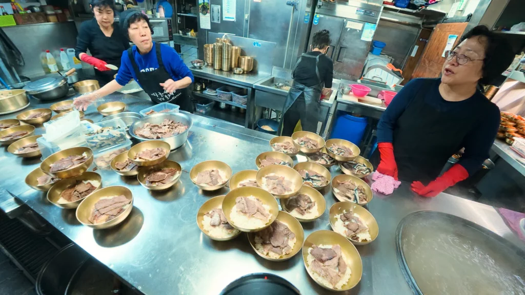 Hadongkwan kitchen staff lining up brass bowls of rice and Hanwoo brisket during the morning gomtang prep rush.