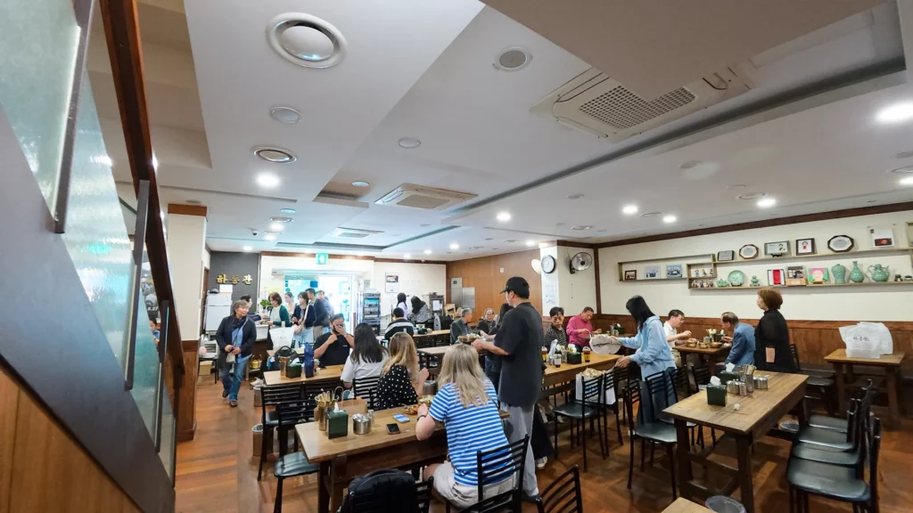 Packed Hadongkwan Myeongdong dining hall during peak hours with diners sharing tables and staff serving brass bowls.
