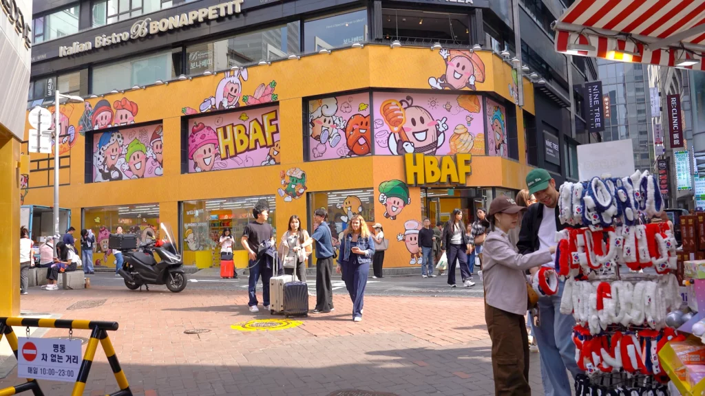 Tourists with suitcases walking past the yellow HBAF Almond Store Myeongdong storefront covered in almond mascot murals.