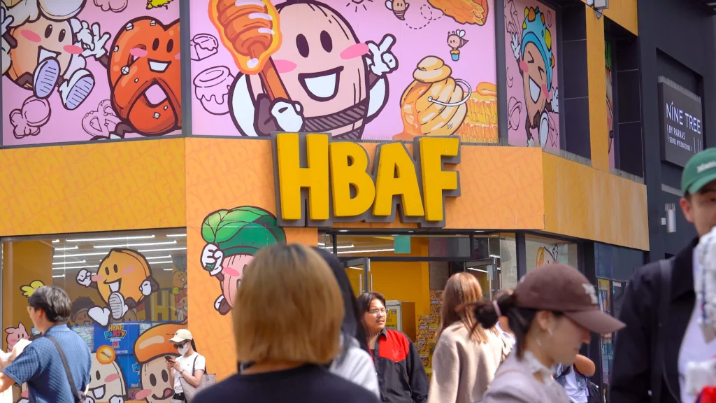 Giant yellow HBAF sign above the entrance of HBAF Almond Store Myeongdong with honey-themed mascot artwork and passing shoppers.
