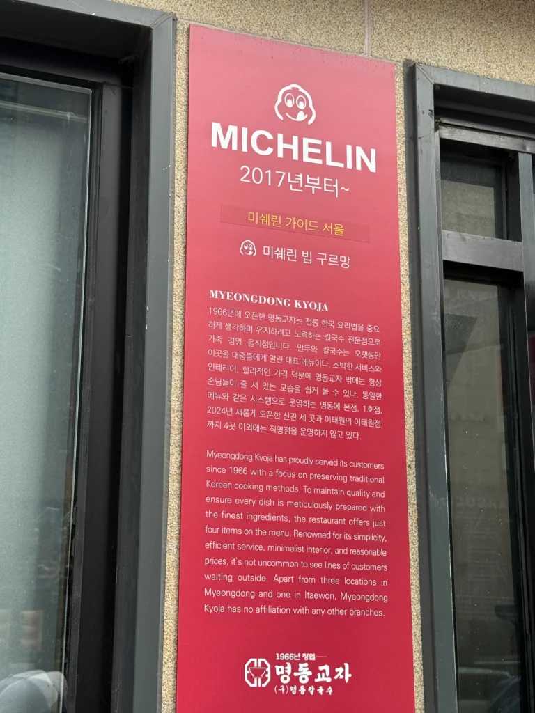 Michelin Bib Gourmand bilingual placard on Myeongdong Kyoja's facade describing the 1966-established kalguksu specialist.