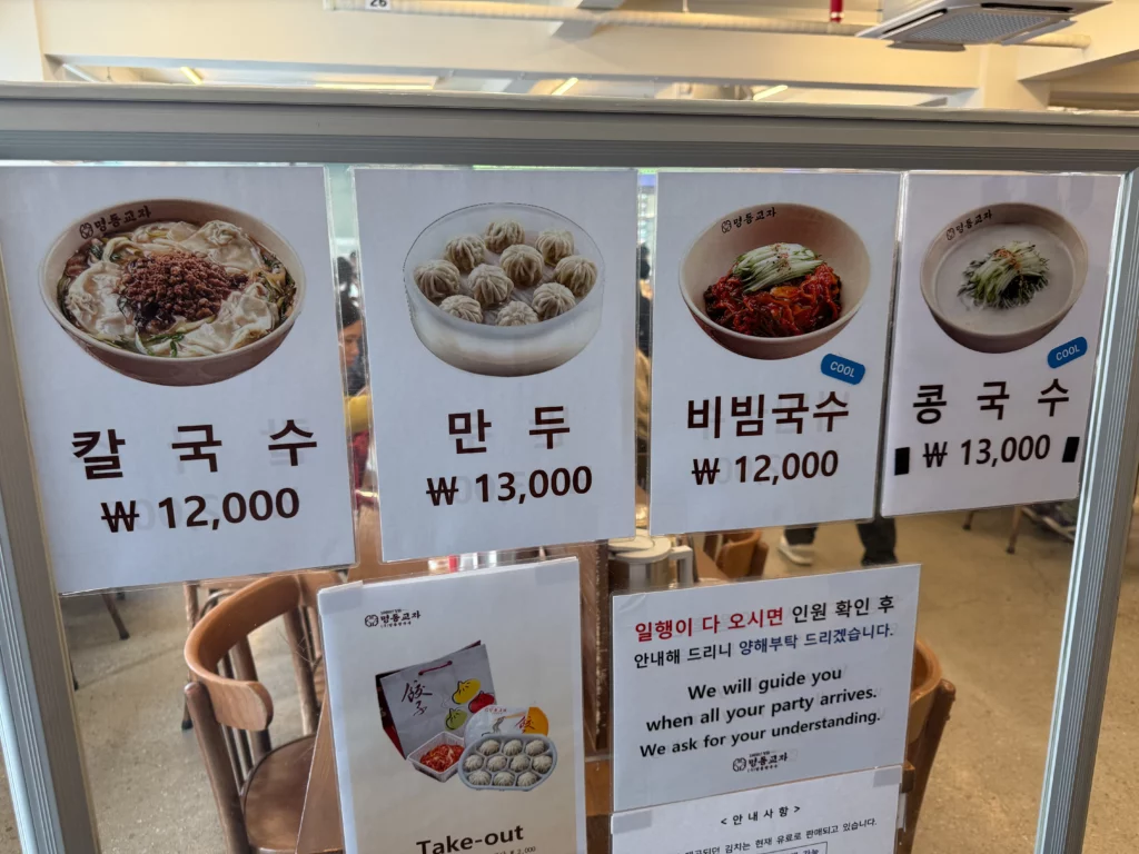 Myeongdong Kyoja menu board listing kalguksu ₩12,000, mandu ₩13,000, bibim-guksu ₩12,000, and kongguksu ₩13,000.
