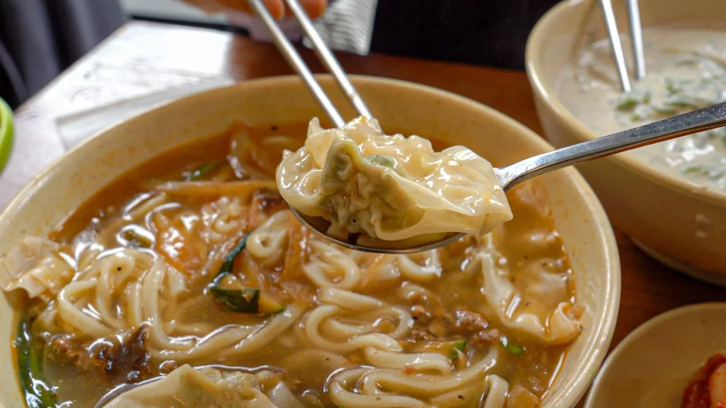 Close-up of a translucent pork-and-chive mandu on a spoon resting on hand-cut kalguksu noodles.
