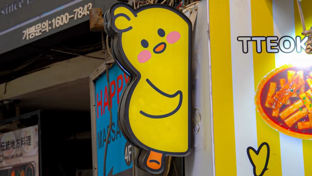 Smiling yellow chick mascot signboard and tteokbokki photo display at the chickgimbap Myeongdong street entrance.