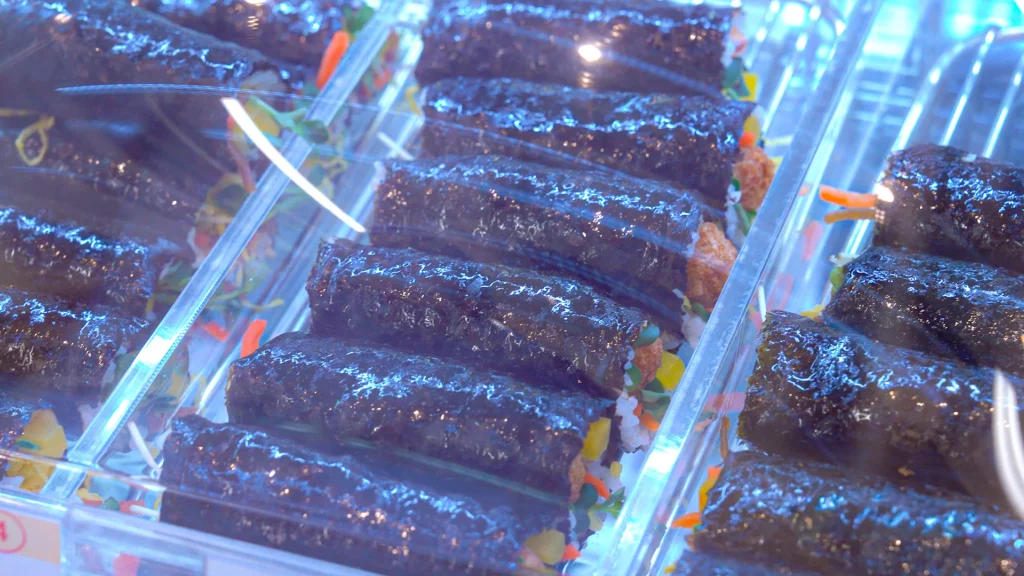 Glossy rows of mini gimbap with colorful vegetable fillings displayed in chickgimbap's brightly lit refrigerated showcase.