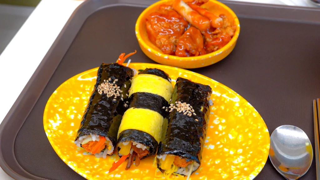 chickgimbap set with three mini gimbap rolls topped with sesame seeds and a side bowl of spicy dak-gangjeong on a yellow tray.