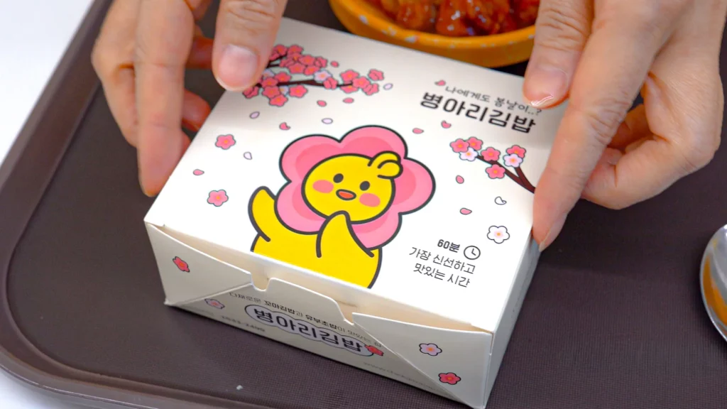 Hands holding a chickgimbap takeout dosirak box decorated with a cherry blossom and Byeongari chick design.