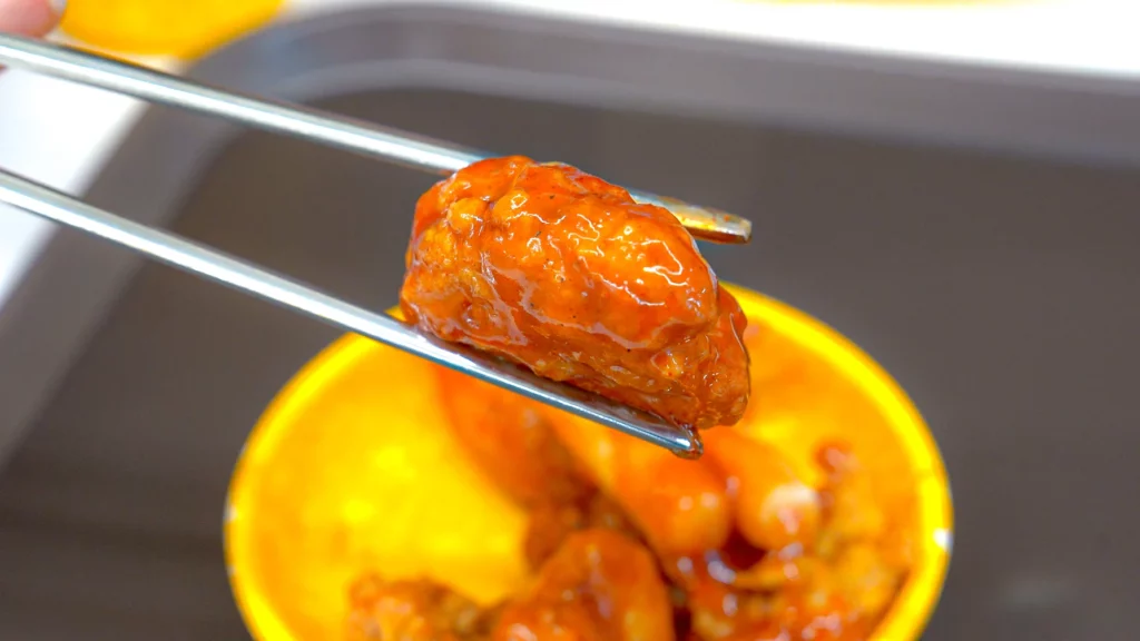 Single piece of glazed Korean dak-gangjeong fried chicken held with metal chopsticks above a yellow bowl.