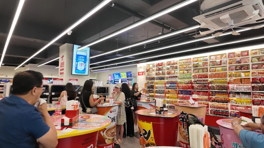 Visitors dining at giant cup noodle-shaped tables inside the CU Hongdae Ramyun Library, with floor-to-ceiling shelves of Korean instant noodles covering the back wall.