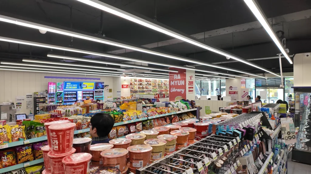 A wide-angle view of the CU Hongdae Ramyun Library interior showing rows of cup ramyun products in the foreground, a full snack aisle, and the Ramyun Library signage and noodle wall visible toward the back of the store.