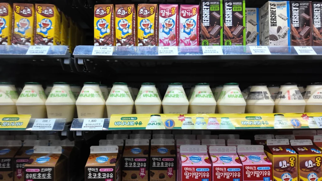 A refrigerated shelf at the CU Hongdae store stocked with Korean flavoured milk drinks including Binggrae 바나나맛 우유 (banana milk), 초코맛 (chocolate), 딸기맛 (strawberry) varieties, and Hershey's chocolate milk cartons — popular beverage pairings with Korean ramyun.