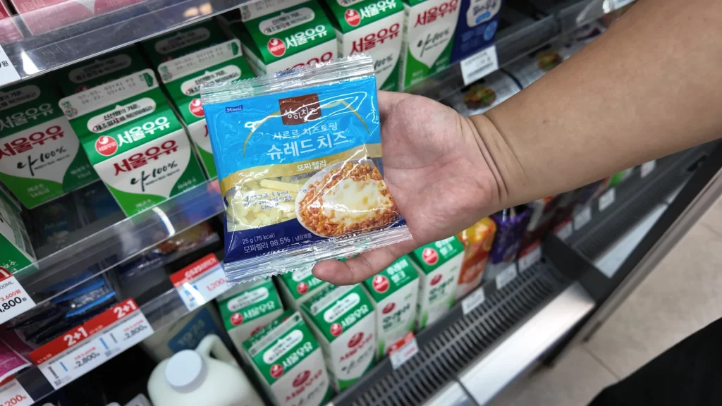 A hand holding a Maeil 쌍치즈 슈레드 치즈 (shredded mozzarella cheese) packet — a popular ramyun topping available at the CU Ramyun Library — in front of a refrigerated dairy section.
