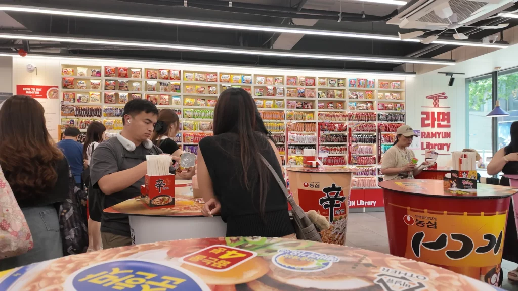 International visitors eating ramyun at Shin Ramyun-branded tables inside the CU Hongdae Ramyun Library, with the full 225-variety noodle wall and 라면 RAMYUN Library logo clearly visible behind them.