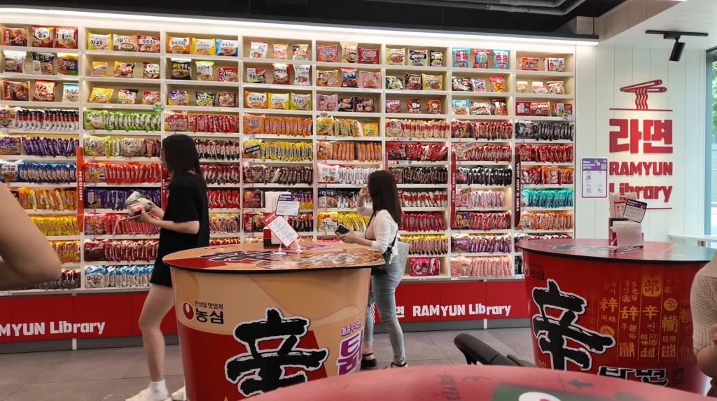 Two visitors browsing the 225-variety ramyun wall at the CU Hongdae Ramyun Library, with the 라면 RAMYUN Library logo displayed on the right wall and Shin Ramyun branded tables in the foreground.