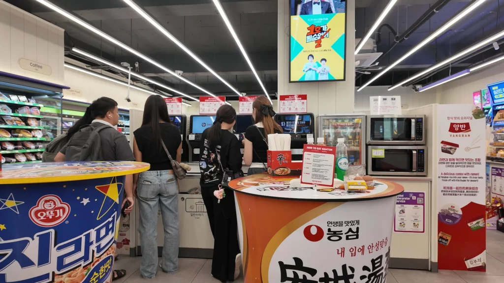 Visitors queuing at the automated cooker stations at the CU Ramyun Library, flanked by Ottogi and Nongshim branded cup noodle-shaped tables, with a How To Eat Ramyun guide displayed on the counter.