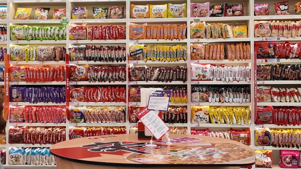 The CU Ramyun Library's floor-to-ceiling instant noodle wall organized by spice level, with Medium and Hot section labels visible and a multilingual translation service card placed on a table.