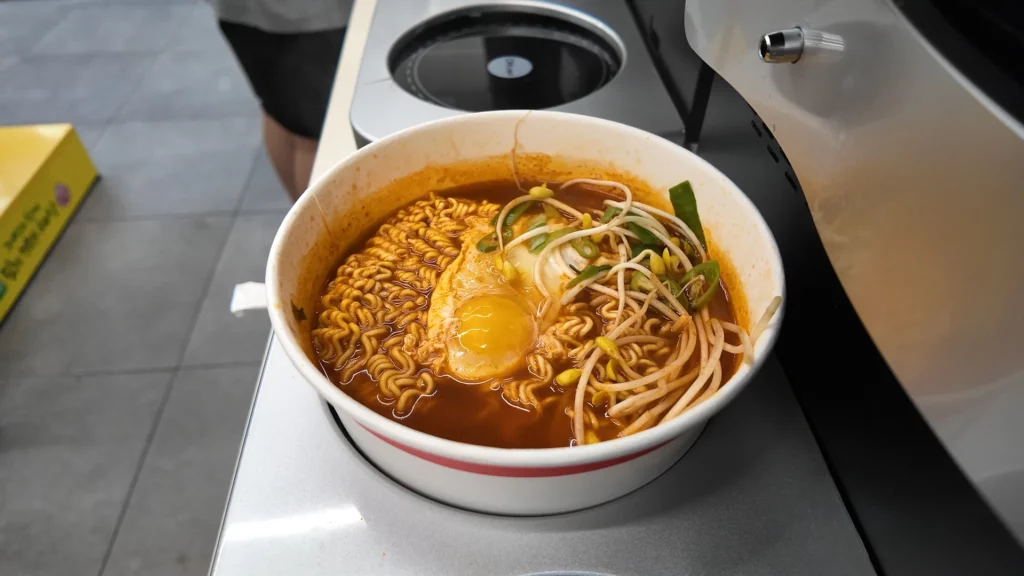 A freshly cooked bowl of spicy Korean ramyun topped with a soft egg, bean sprouts, and green onions, prepared at the CU Ramyun Library's automated cooker station.