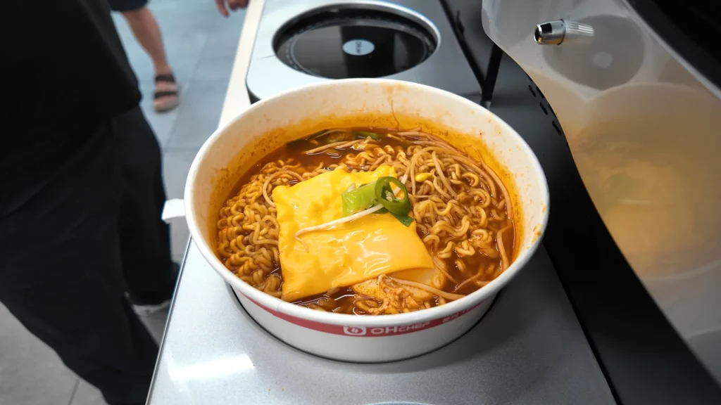 A bowl of Korean ramyun topped with a melted processed cheese slice and sliced green chili, prepared at the CU Ramyun Library's automated cooker — showcasing the popular cheese topping combination.