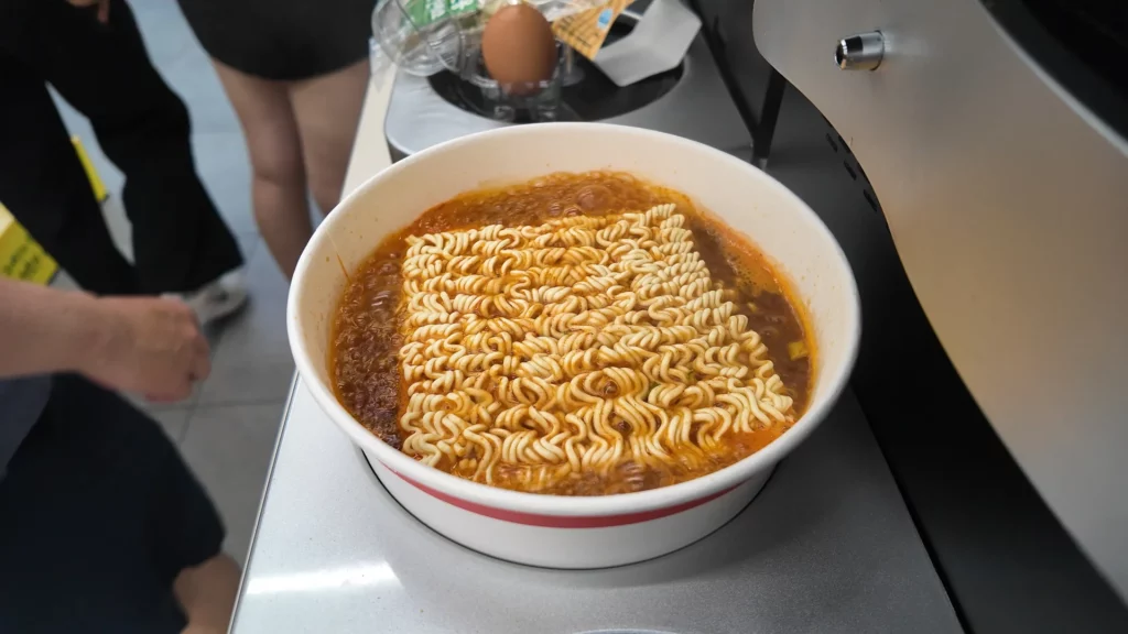 A paper cooking bowl filled with freshly dispensed spicy ramyun broth and wavy noodles sitting on the automated cooker station at the CU Ramyun Library, just after cooking.
