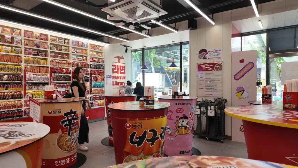 A wide interior view of the CU Hongdae Ramyun Library showing the full ramyun wall, multiple giant cup noodle-shaped dining tables branded with Shin Ramyun, Neoguri, and Buldak, and large windows overlooking the Hongdae street outside.