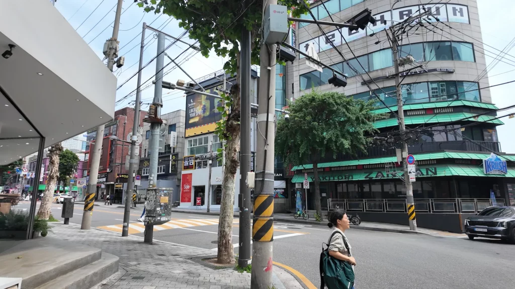 A street-level view of the Hongdae neighbourhood in Seoul near the CU Ramyun Library, showing a busy intersection lined with shops and commercial buildings on a clear day.