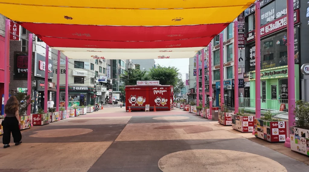 The colourful pedestrian plaza in the Hongdae neighbourhood near the CU Ramyun Library, featuring a red and yellow canopy, pink columns, and a ReoReo-branded street performance stage at the far end.